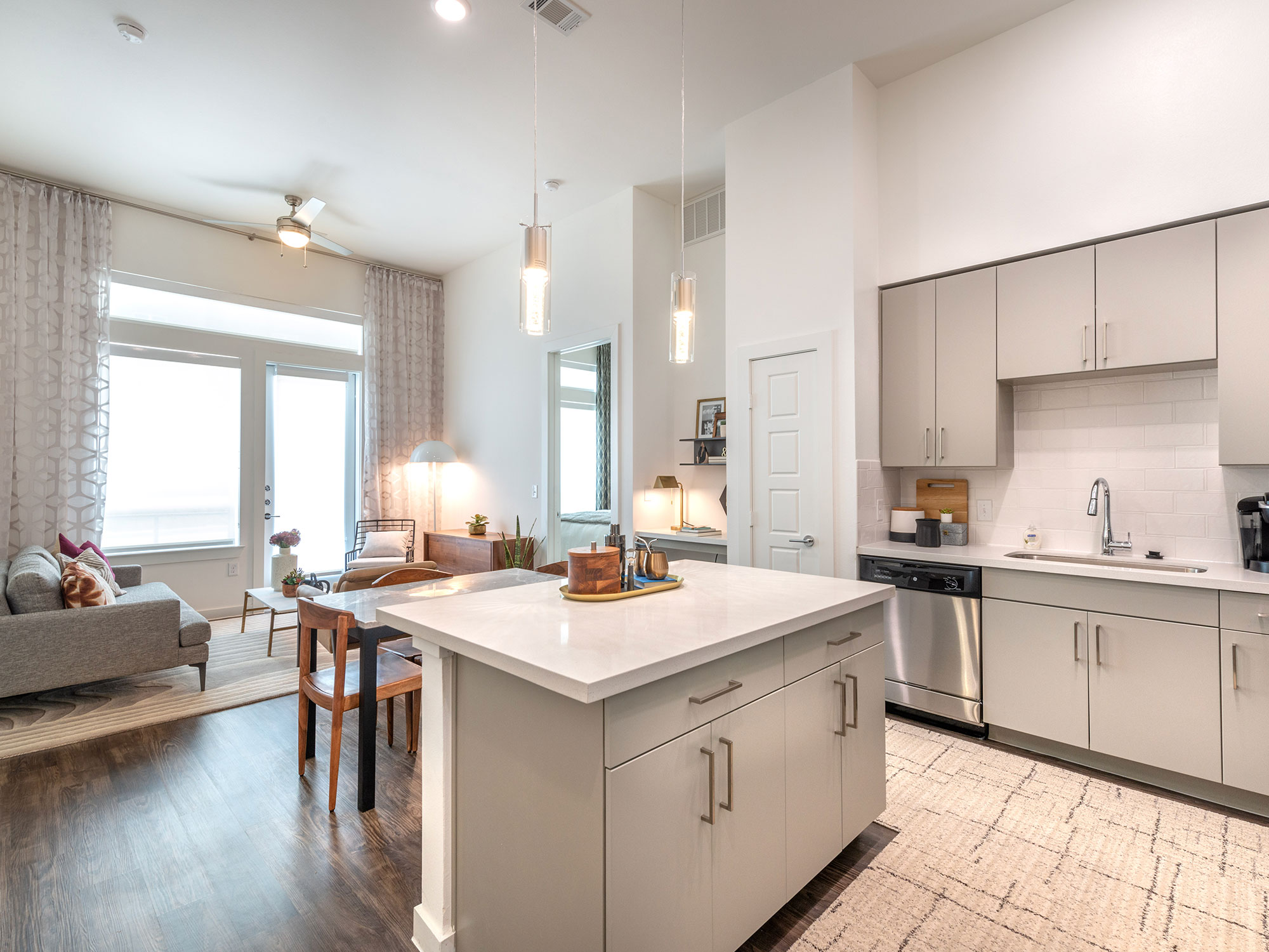 Apartment interior with plank flooring, modern kitchen with stainless steel appliances and designer lighting, and living room with built-in desk and a door leading to patio