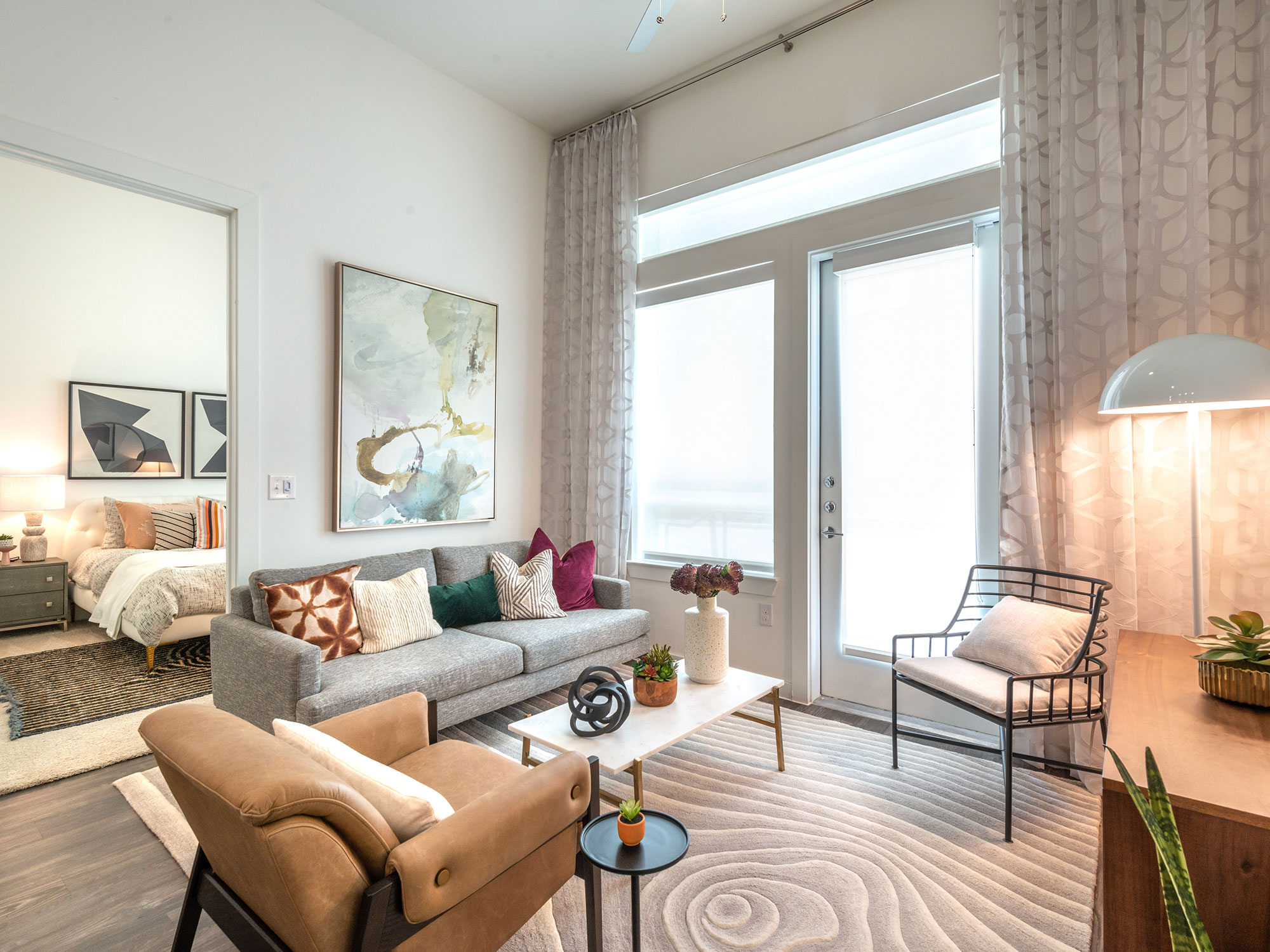 Living room with ceiling fan, plank flooring, large windows, entrance to bedroom and door to the patio