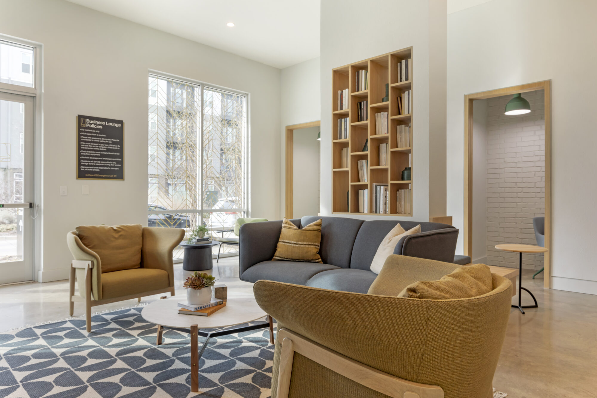 Modern lounge with a gray sofa, mustard yellow armchairs, round coffee table, geometric rug, bookshelves, large window, and neutral decor. Bright, inviting atmosphere with natural light.